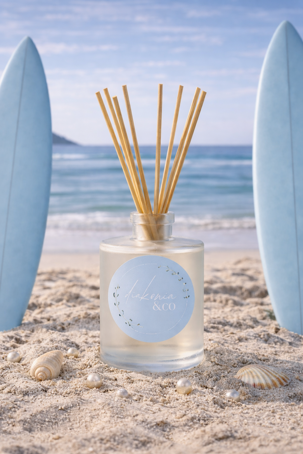 Reed diffuser with wooden sticks in a glass bottle on a sandy beach with surfboards and ocean in the background.