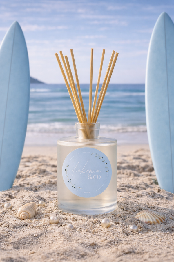 Reed diffuser with wooden sticks in a glass bottle on a sandy beach with surfboards and ocean in the background.