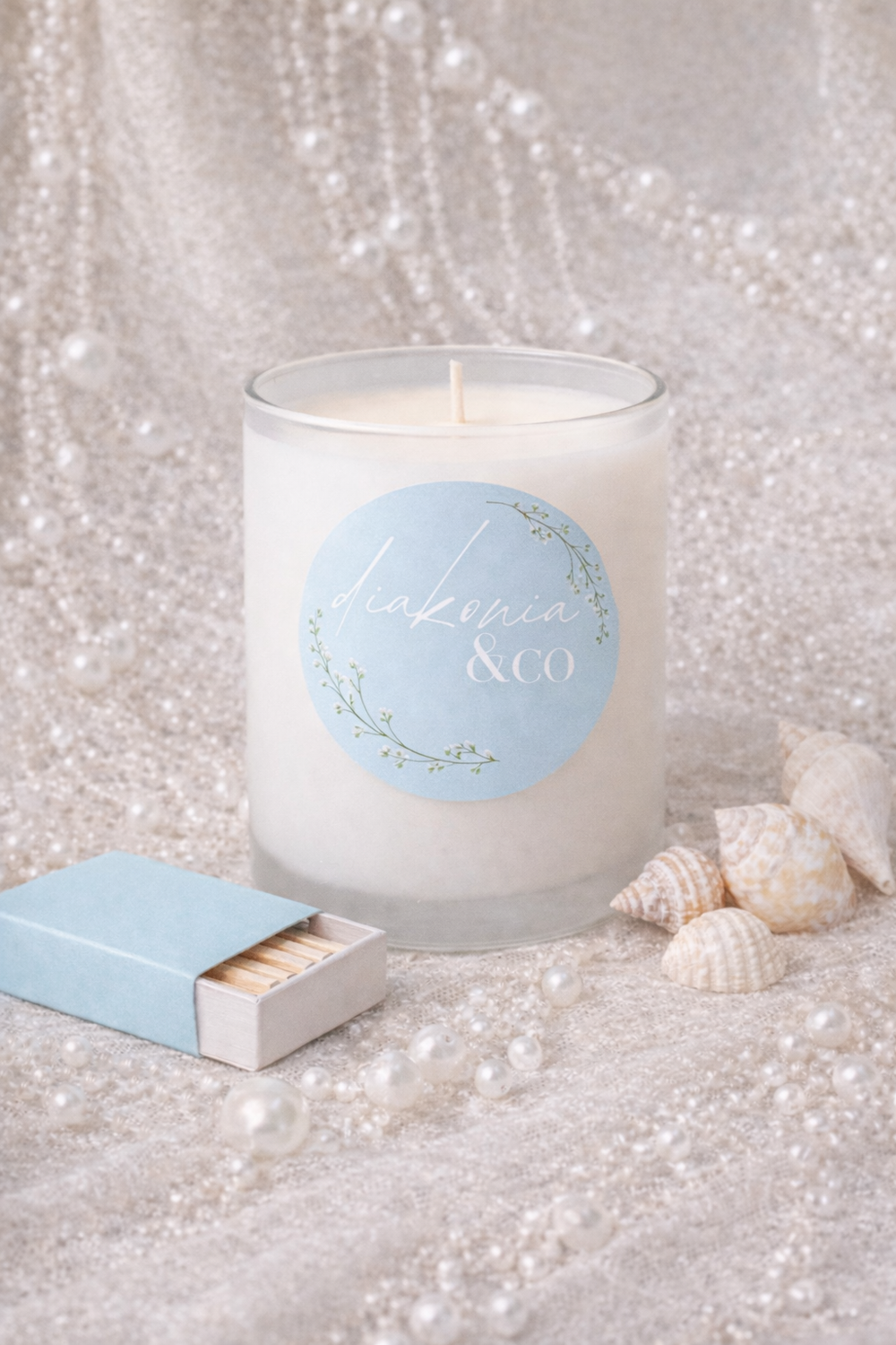 Candle with a blue label and matchbox on a textured surface with pearls and shells.